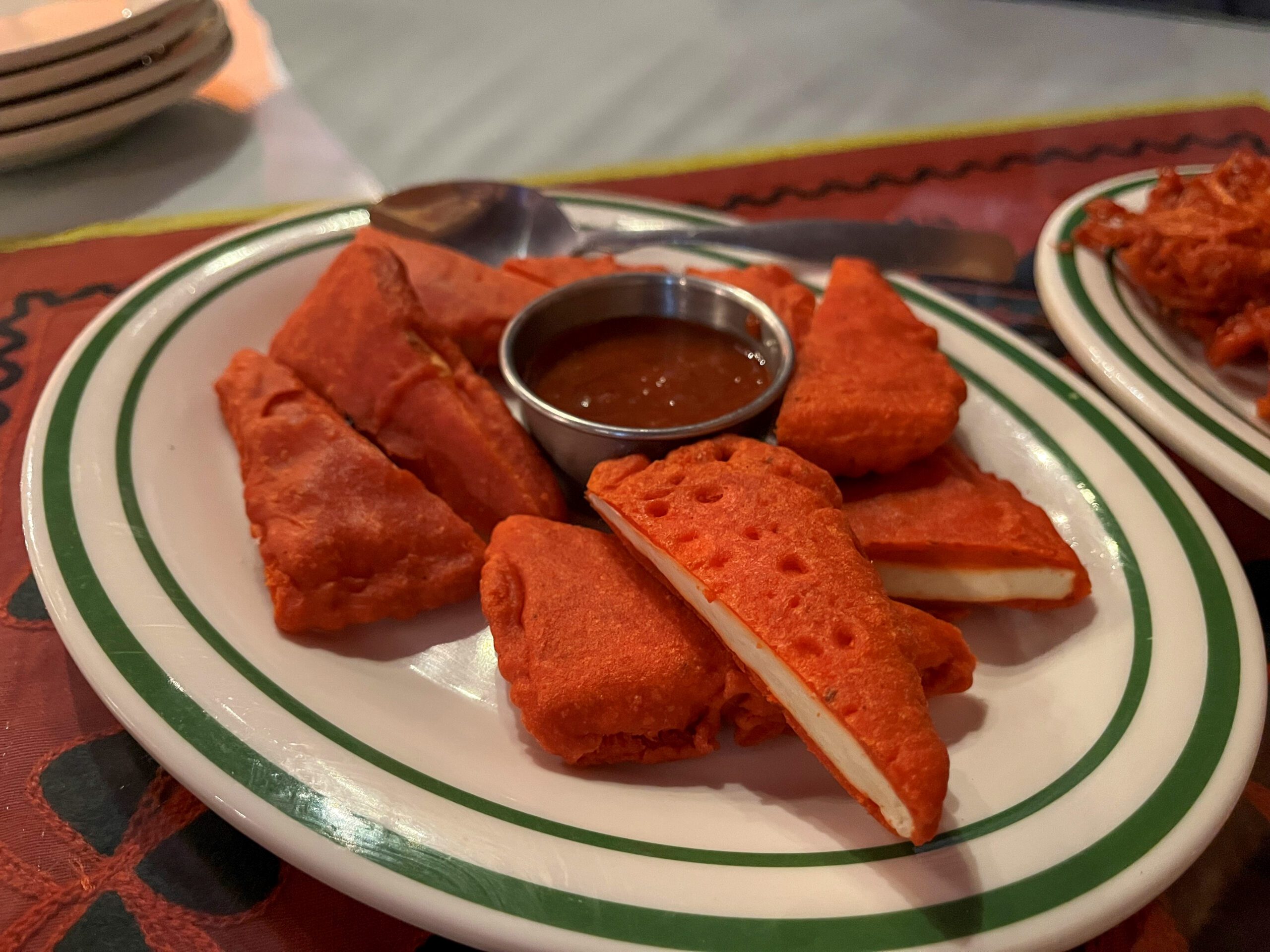 A plate with several pieces of deep-fried, orange-colored paneer pakora arranged around a small metal cup filled with dipping sauce. The pakoras are crisp and golden, with visible slices of white paneer inside. The table is set on a colorful red tablecloth.
