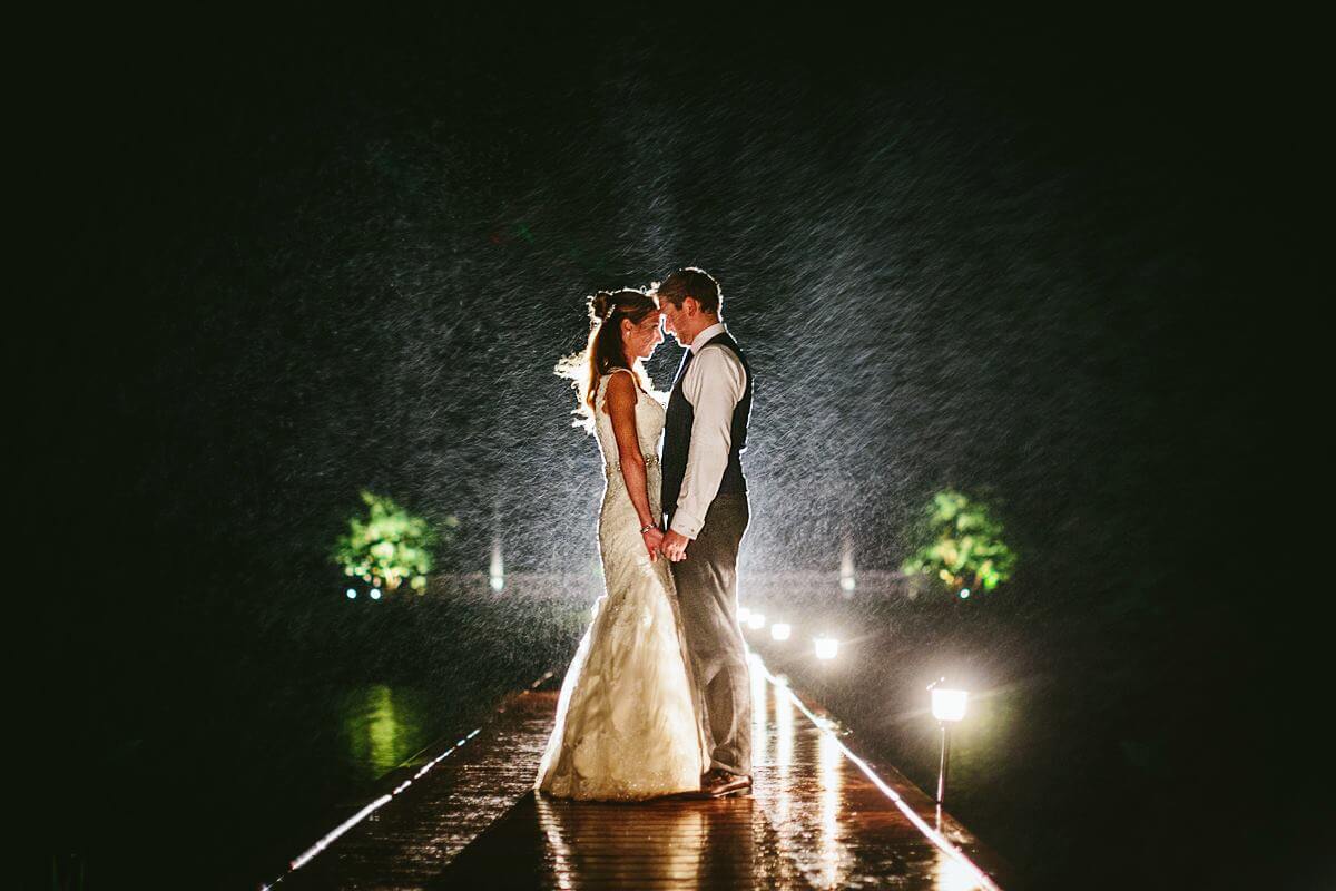 A bride and groom stand face to face, holding hands on a wet, illuminated dock at night. The bride is in a sleeveless white gown, and the groom is in a vest and tie. They are surrounded by falling rain with greenery faintly visible in the background.