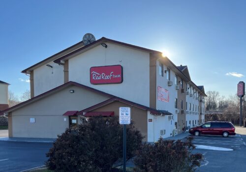 A Red Roof Inn building with beige exterior walls and a red roof. The hotel has two visible signs with the Red Roof Inn logo. A red van is parked in the hotel's parking lot next to a shrubbery. The sky is clear, and the sun is low, casting long shadows. Image