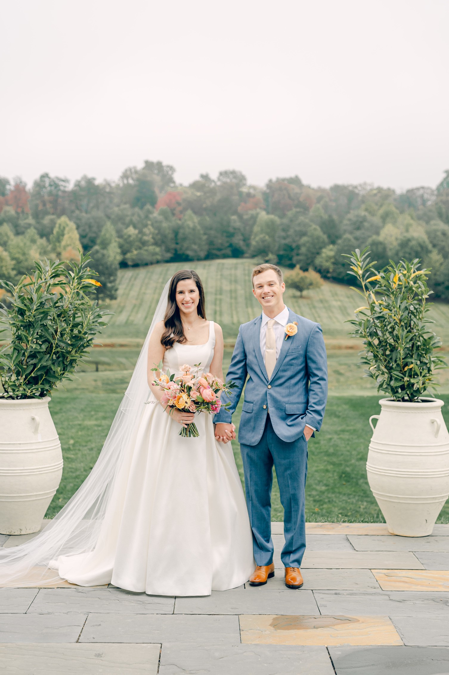 A bride in a white wedding dress holding a bouquet and a groom in a blue suit with a boutonnière stand hand in hand outside on a stone pathway. They are flanked by large white planters with greenery. A lush green lawn and trees are visible in the background.
