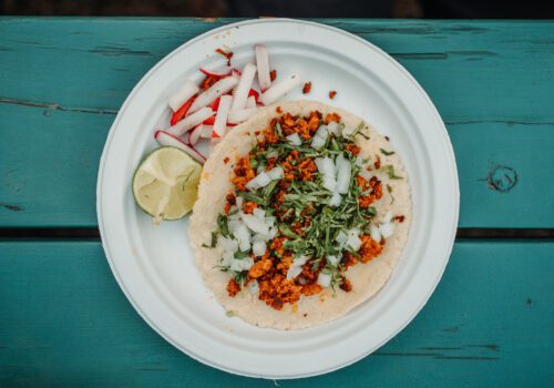 A white paper plate with a taco filled with meat, chopped onions, and cilantro sits on a turquoise wooden table. The plate also contains sliced radishes and a lime wedge on the side. Image