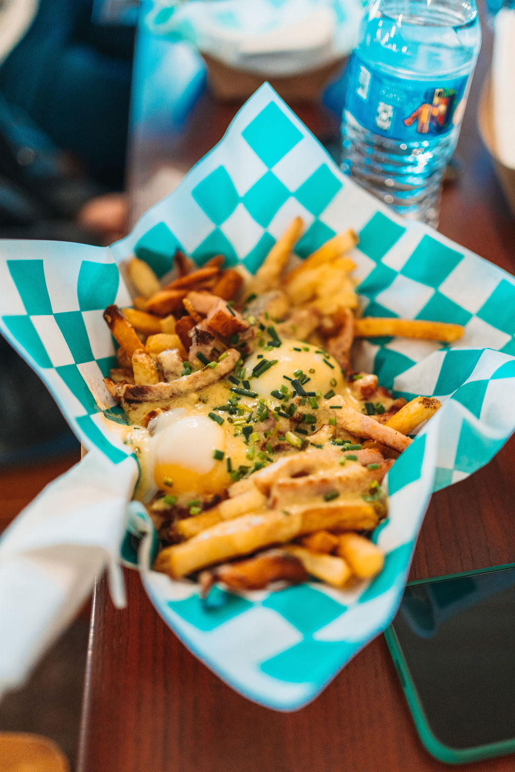 A basket lined with blue and white checkered paper holds a serving of fries topped with melted cheese, two eggs, chives, and bacon bits. There is a water bottle and a smartphone on the table beside the basket.