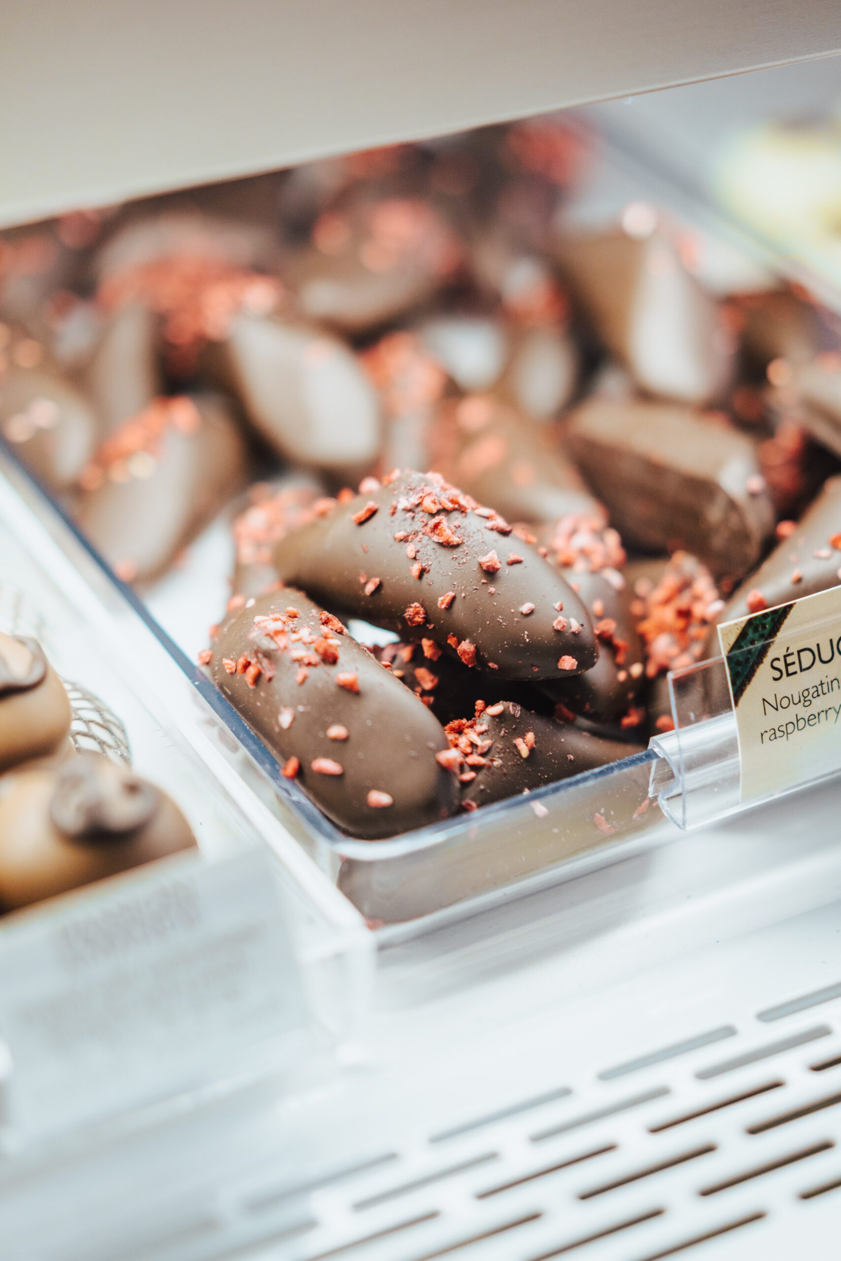 Close-up of a display case showcasing chocolate-covered treats sprinkled with dried raspberry pieces. The shelf label partially visible nearby mentions 