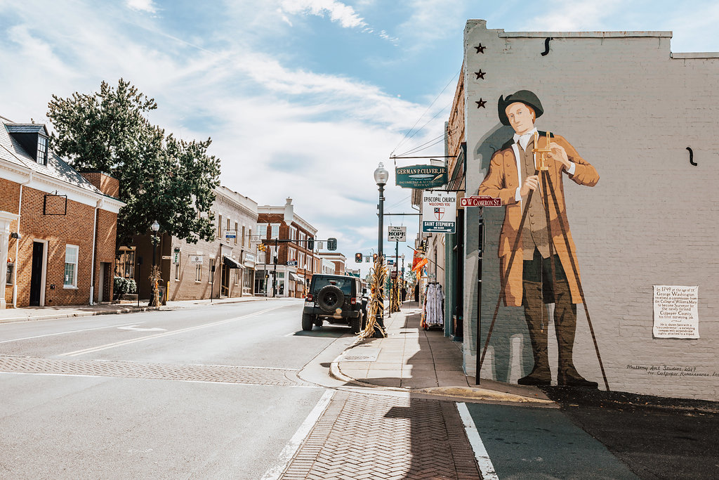 A quiet street in a small town featuring a large mural of a historic figure holding a telescope painted on the side of a building. The street is lined with shops, a lamppost with directional signs, and a jeep is parked along the sidewalk. The sky is partly cloudy.