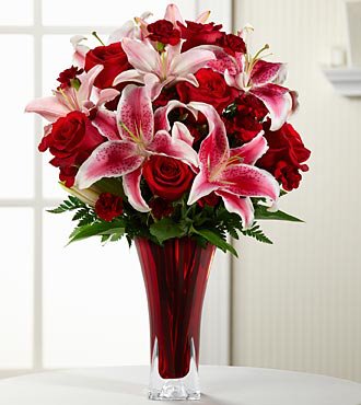A bouquet of pink stargazer lilies and red roses arranged in a tall, elegant red glass vase. The flowers are surrounded by green foliage and are displayed in a well-lit room with a light-colored background.