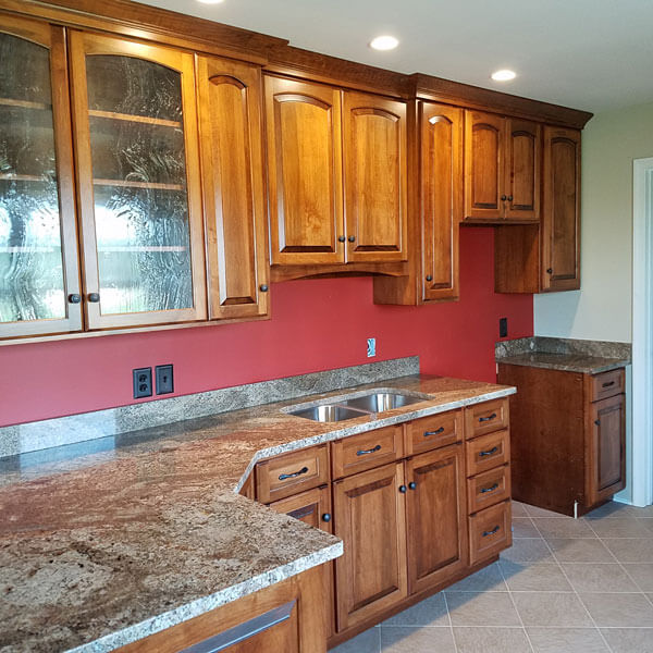 A kitchen with wooden cabinets and brown marble countertops. The cabinets have glass fronts on some doors, and there is a double sink below one. The backsplash and wall are painted red. The floor is covered with beige tiles. Recessed lighting is installed in the ceiling.