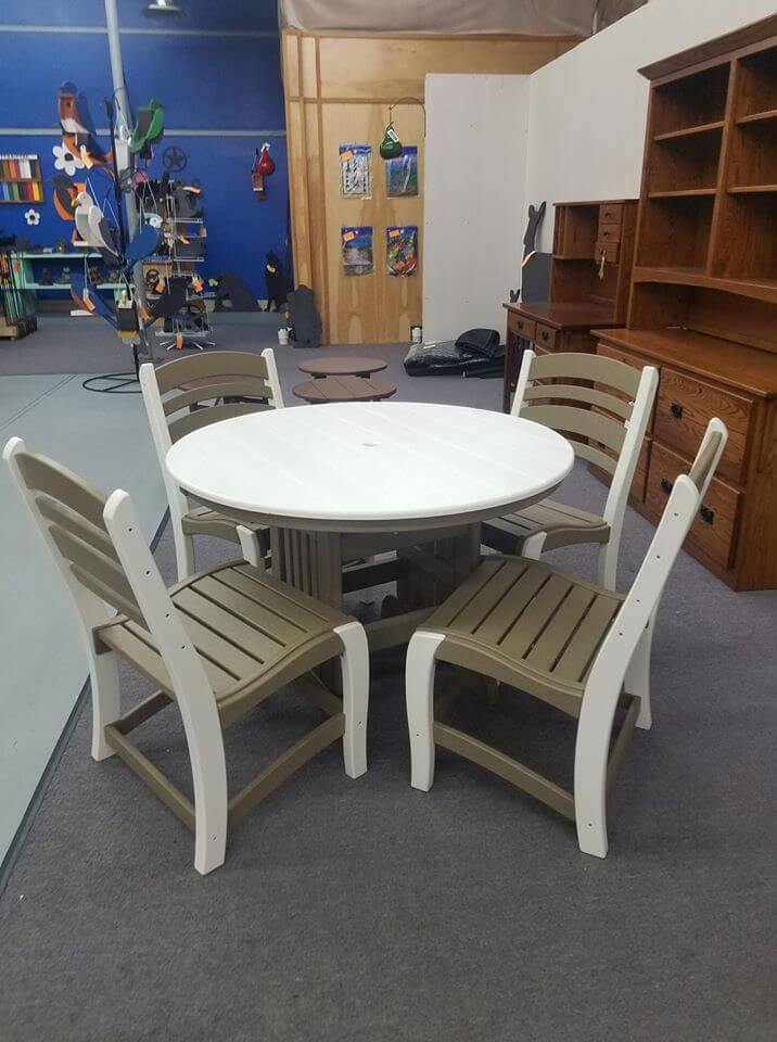A round white table with a pedestal base is surrounded by four chairs with white frames and beige seats and backs. Various wooden furniture pieces and shelves are visible in the background, along with a colorful, abstract wall decoration.