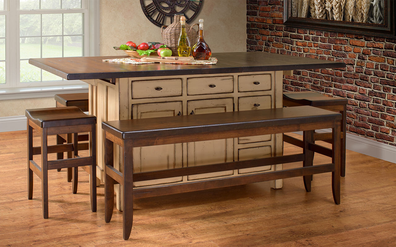 A rustic kitchen island with a wooden countertop stands against a brick wall. Four wooden stools are placed around it. The island is adorned with fresh produce, olive oil, a glass container, and decorative items. Sunlight filters in through a nearby window.