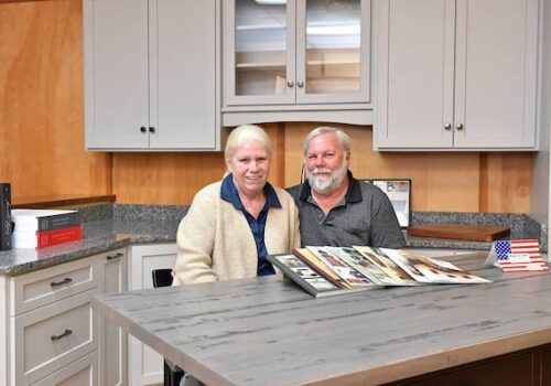 An older man and woman are seated at a kitchen island, looking at a photo album. The woman wears a light-colored sweater and the man has a gray beard and wears a dark shirt. Behind them, there are cabinets, counters, and a small American flag on the kitchen island. Image
