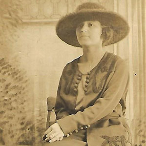 A sepia-toned photograph of a woman wearing a wide-brimmed hat and an ornate buttoned jacket. She is seated on a chair, looking slightly to the side, with her hands gently clasped in her lap. The backdrop includes subtle, intricate patterns and foliage.