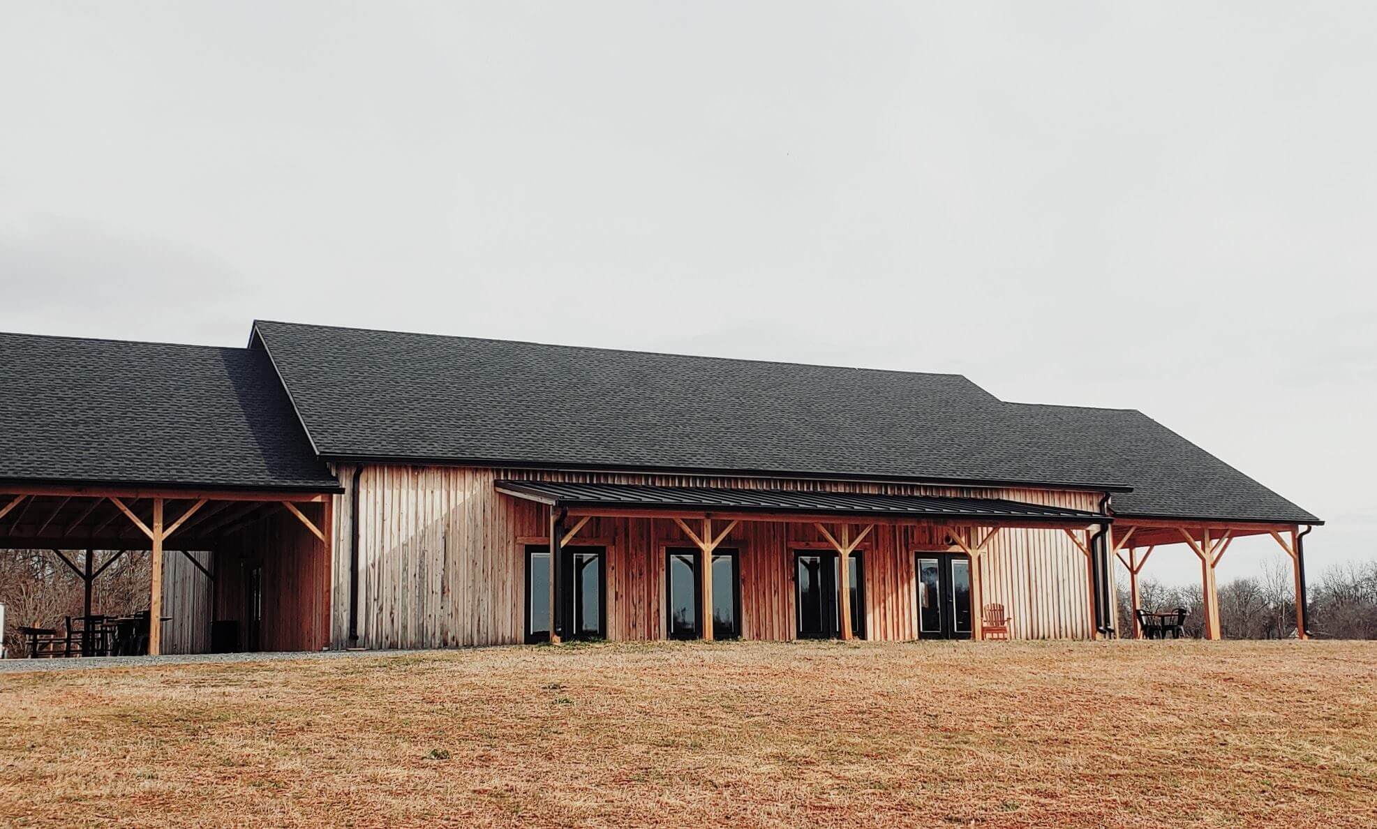 A large wooden building with a dark roof sits on a grassy area with scattered brown patches. The structure has numerous large glass doors and windows, and a covered outdoor space on the left side. The sky overhead is cloudy.