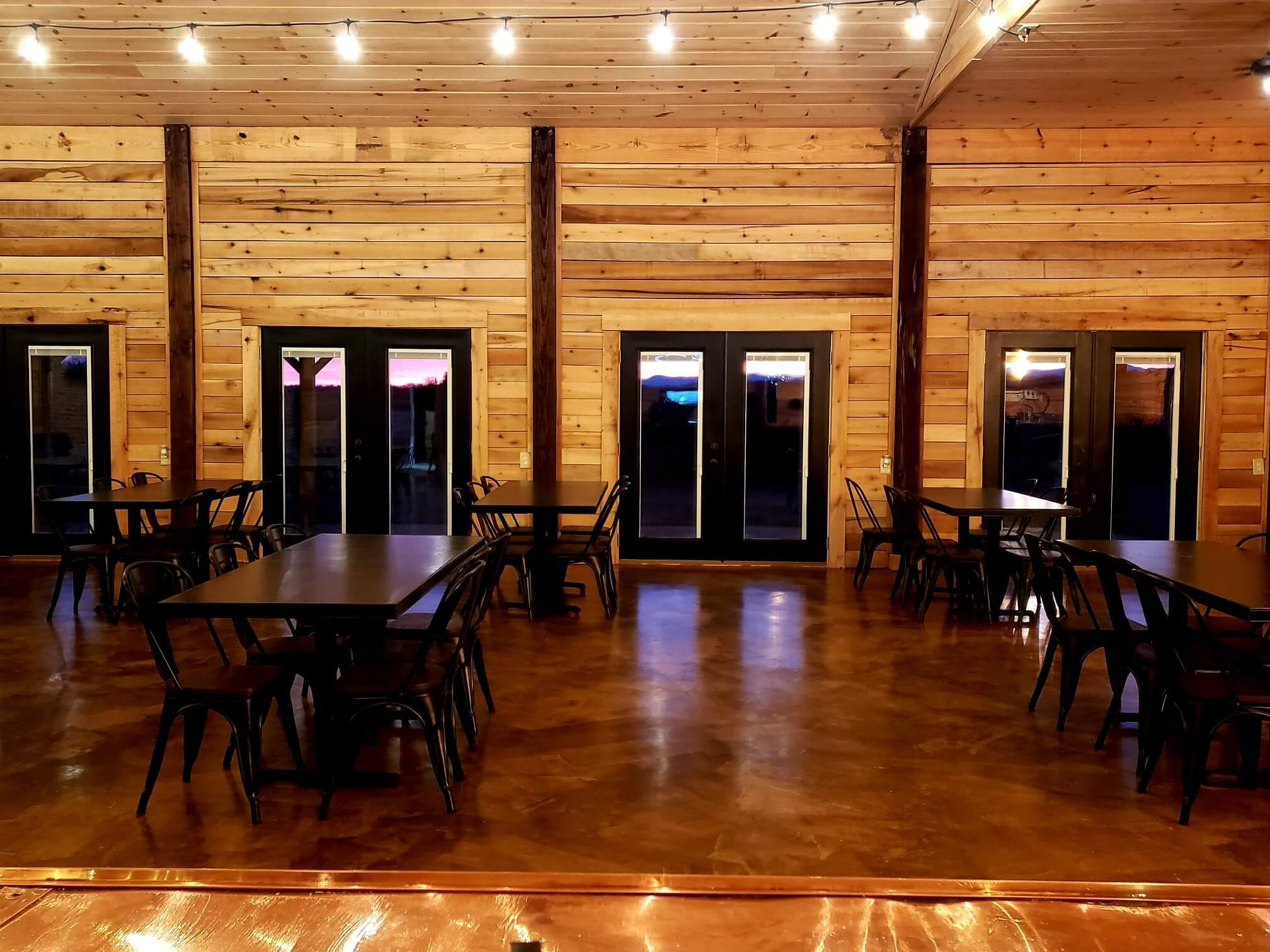 A warmly lit room with wooden walls, ceiling, and floor features several black tables and matching chairs. String lights run along the ceiling, and large windows and doors line the back wall, revealing a sunset outside. The room has a rustic, cozy ambiance.