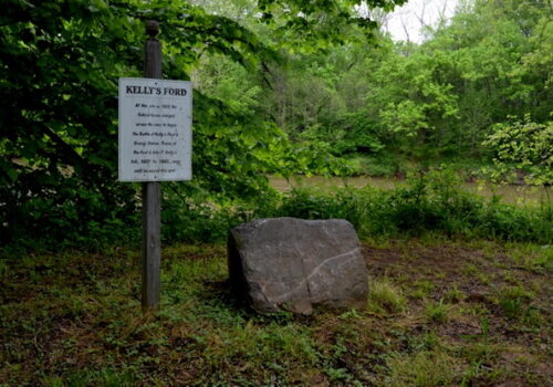 A large rock and a signpost stand in a green, leafy area next to a river. The sign reads 