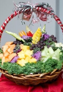 A festive wicker basket filled with an assortment of fruits and vegetables, including pineapple, melon, grapes, carrots, and kale. The basket is adorned with a red and white striped handle and topped with a decorative bow in matching colors. Image