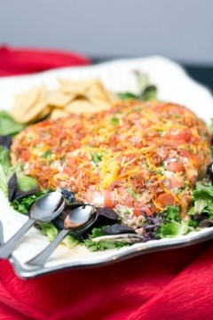 A heart-shaped dish filled with a colorful chopped salad, topped with grated cheese, tomatoes, and green herbs. The platter rests on a bed of mixed greens and is accompanied by two spoons and a small pile of tortilla chips. The background features a red cloth.