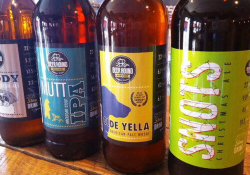 A close-up of four beer bottles lined up on a wooden surface. The labels read 