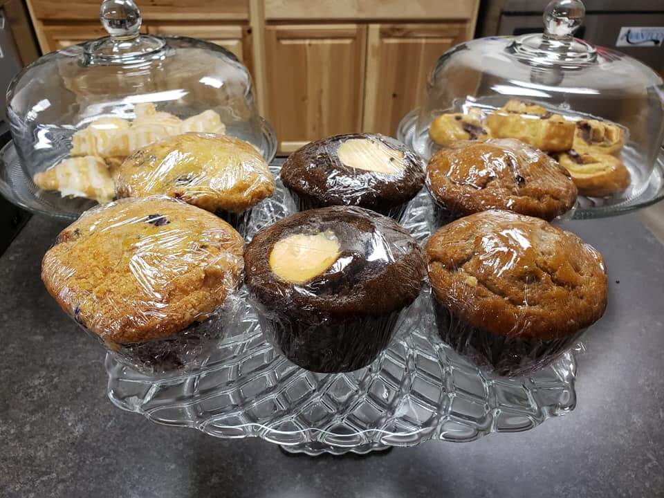 A glass display stand holds various individually wrapped muffins and pastries. There's a mix of blueberry, chocolate, and walnut muffins in the foreground, while two glass domes in the background showcase danishes and pastries, all set on a dark countertop.