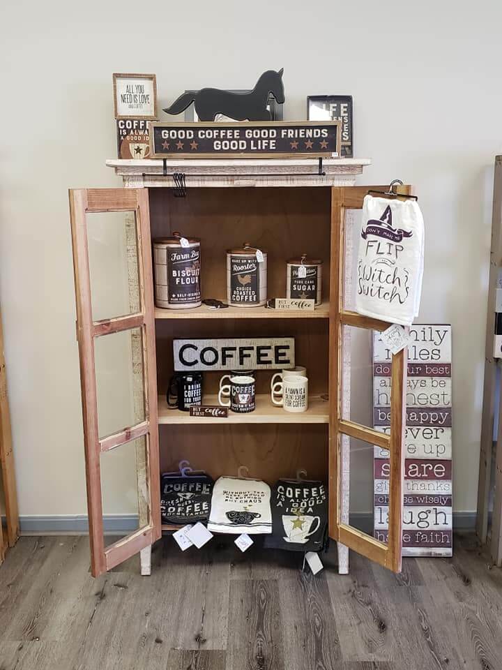 A wooden display cabinet with glass doors showcases various coffee-themed kitchen decor items. The shelves hold mugs, signs, and containers all related to coffee. The surrounding area has additional decorations, including a sign with positive affirmations.