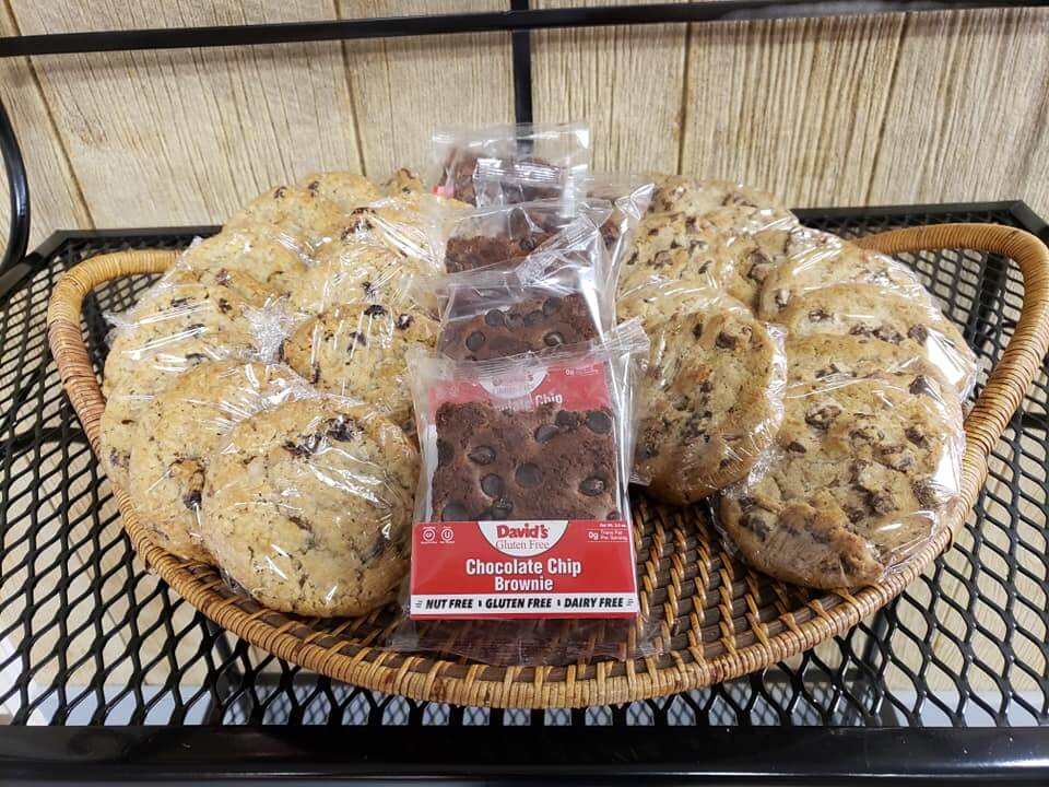 A wicker tray on a black metal shelf holds individually wrapped cookies and packages of brownies. The cookies are chocolate chip, and the brownies are labeled 
