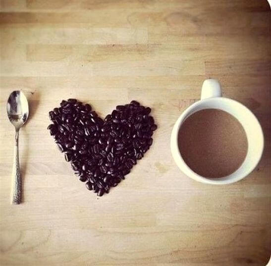 A spoon, a group of coffee beans arranged in the shape of a heart, and a white mug filled with coffee are placed side by side on a wooden surface.