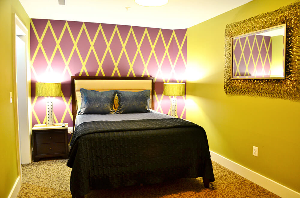 A bedroom with a green and purple diamond-patterned accent wall behind a bed with navy bedding. The bed is flanked by two nightstands with green lamps. The room features green walls, a gold-framed mirror, and a beige carpet.