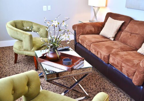 A cozy living room with a brown sofa adorned with patterned cushions, two green armchairs, and a glass coffee table. The coffee table holds a decorative tray with a vase containing white flowers and a small stack of coasters. A floor lamp stands in the corner. Image