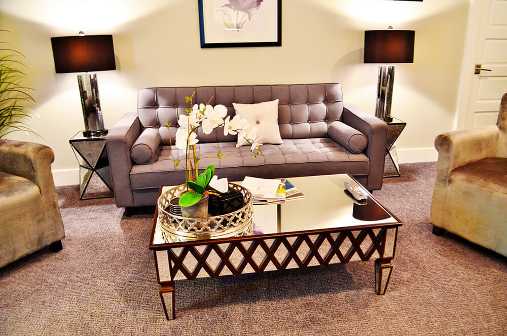 A cozy living room features a tufted gray sofa with decorative pillows, flanked by modern black lamps on side tables. A glass-top coffee table with a decorative tray and orchid centerpiece sits in front, flanked by two soft, gray armchairs. Beige carpet covers the floor.