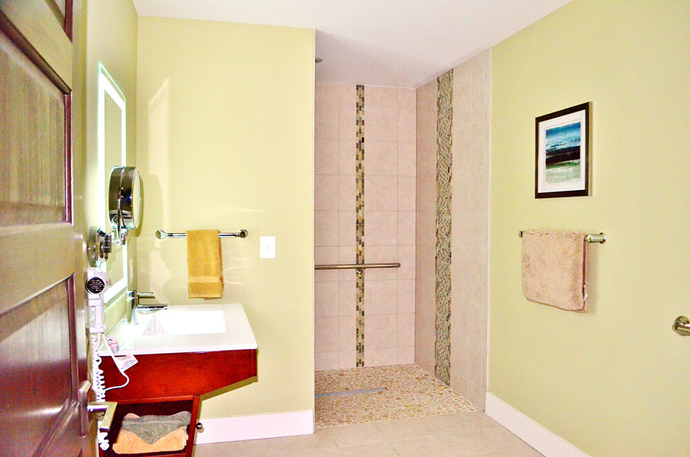 A modern bathroom features light green walls, a walk-in shower with mosaic tile accents, a white sink with a brown vanity, a round mirror, a towel rack with yellow and beige towels, and wall art with a landscape painting. The floor and shower are tiled.