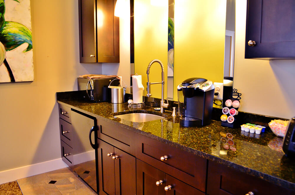 A modern kitchenette with dark wood cabinets and a polished granite countertop. There is a stainless steel sink with a tall faucet, a coffeemaker, a variety of coffee pods, and a microwave. Bright lighting and a partial view of a decorative painting are visible.