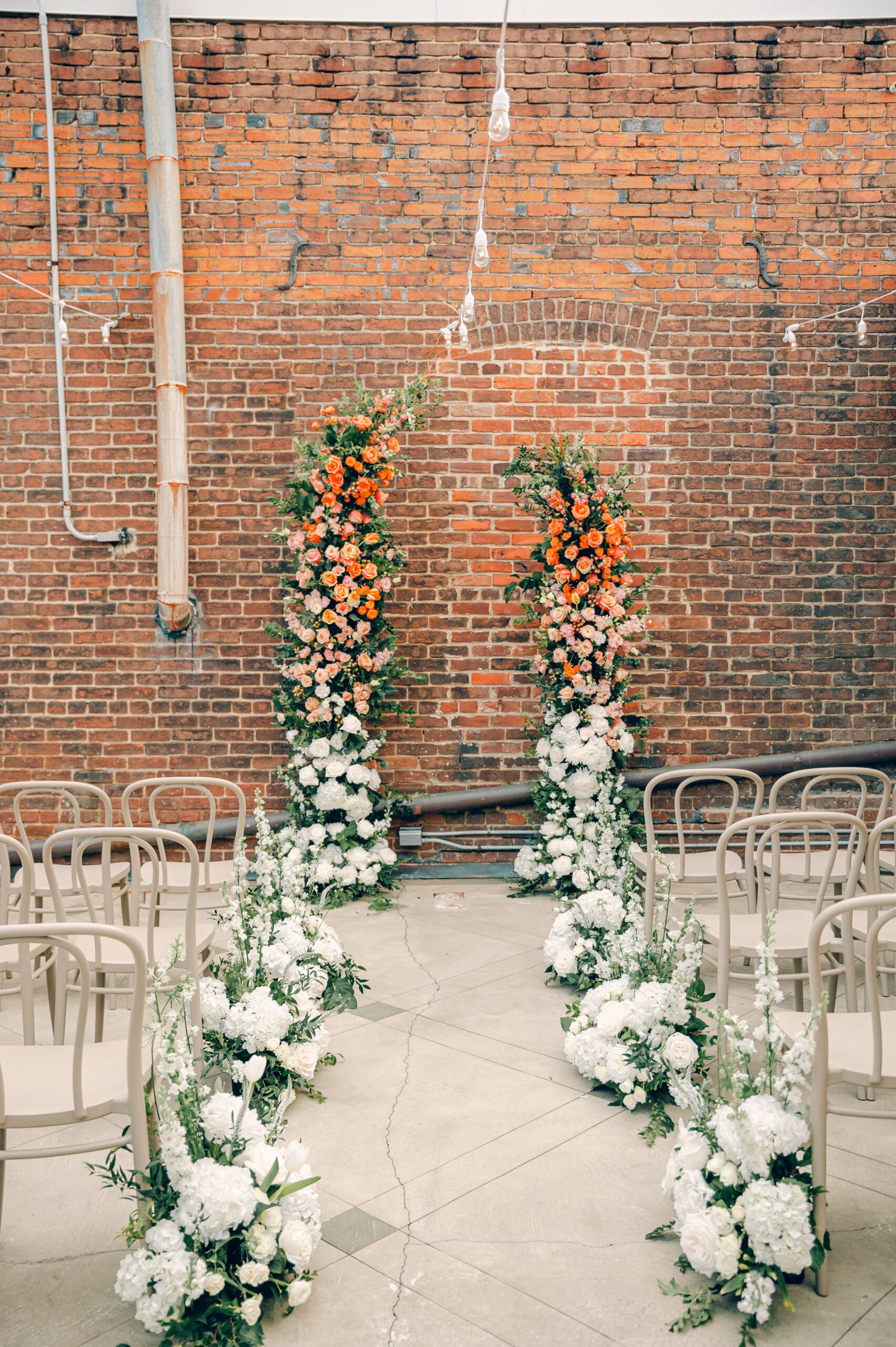 A wedding ceremony setup with two tall floral arrangements featuring orange and white flowers against a brick wall. White flowers line the aisle, which is surrounded by beige chairs. Strings of lights hang above, creating a cozy atmosphere.