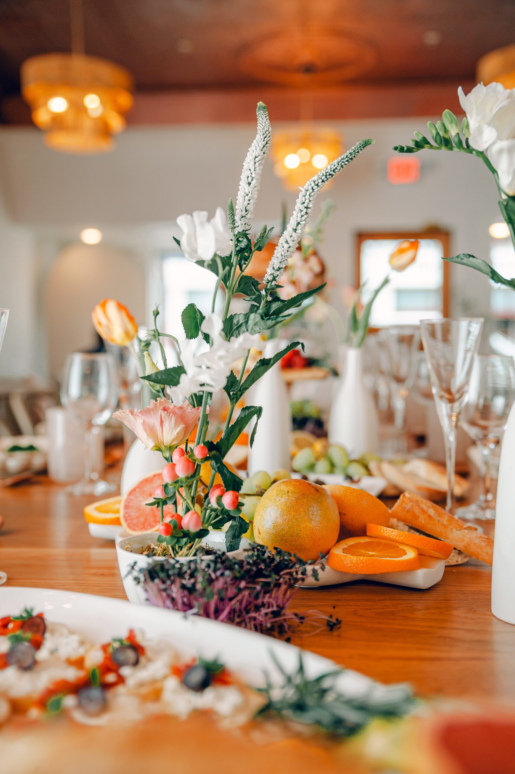 A beautifully set dining table featuring elegant floral centerpieces, sliced fruits including oranges and grapefruit, and an assortment of appetizers. Wine glasses are placed at each setting, and the background shows a warmly lit, sophisticated dining area.