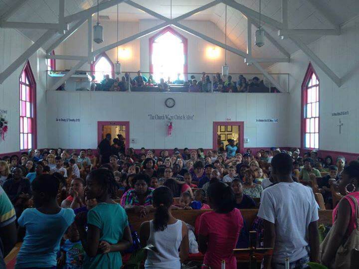 A large gathering of people inside a church, filling both the main floor and the balcony. The attendees, including many children, are seated facing the front. The interior features white walls, red-trimmed windows, and a high ceiling.