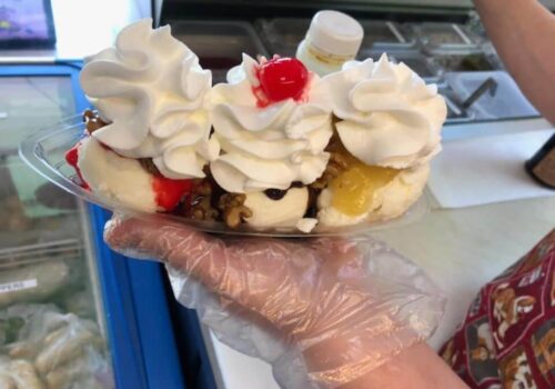 A person wearing a glove holds a boat-shaped dish with three scoops of ice cream, each topped with whipped cream, a cherry, and assorted toppings like nuts and syrup. The background shows a counter with various containers, and part of the person's arm is visible. Image