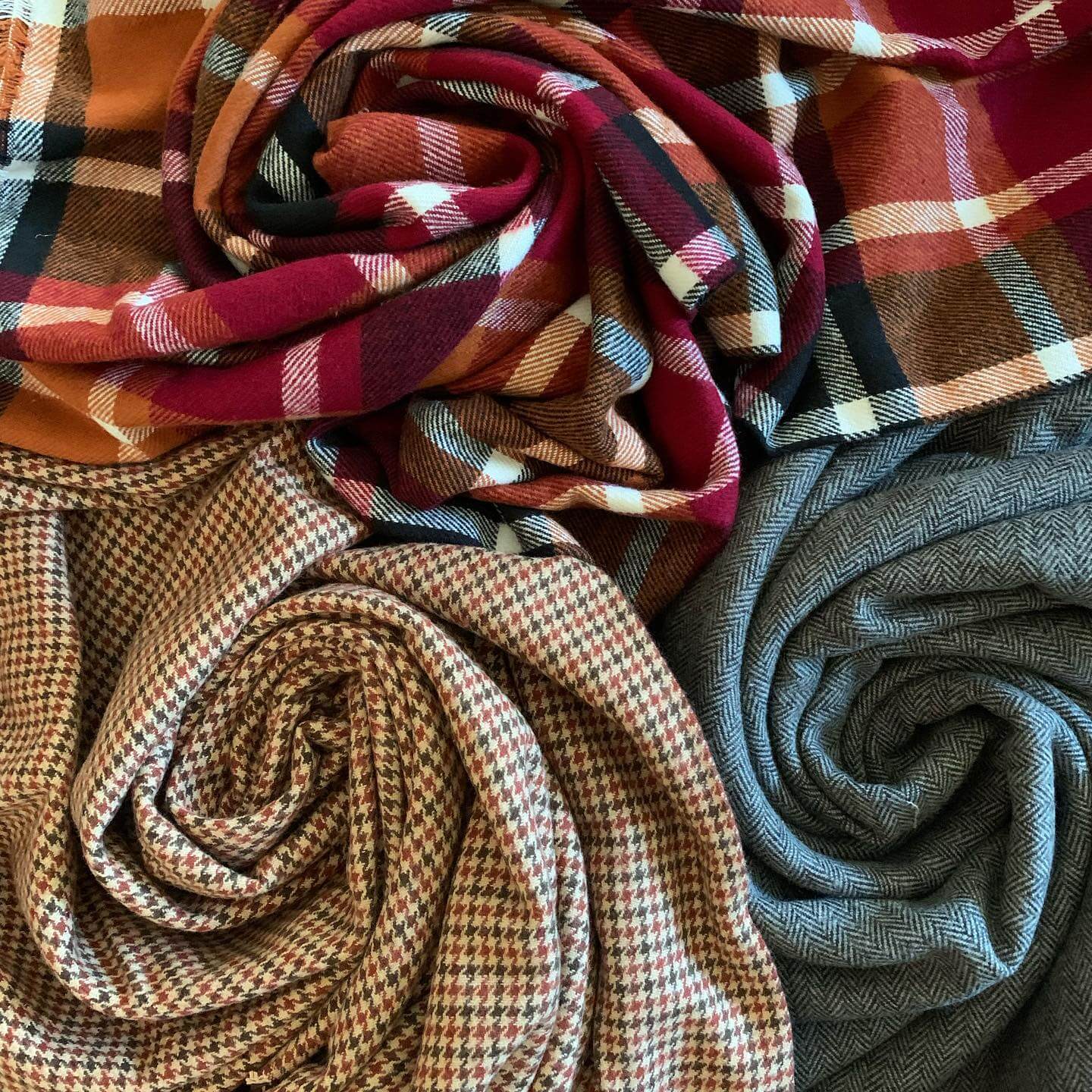 A close-up image of three types of fabric swirled together: a red, black, and white plaid on top, a brown and black houndstooth in the middle, and a grey with a subtle herringbone pattern at the bottom.