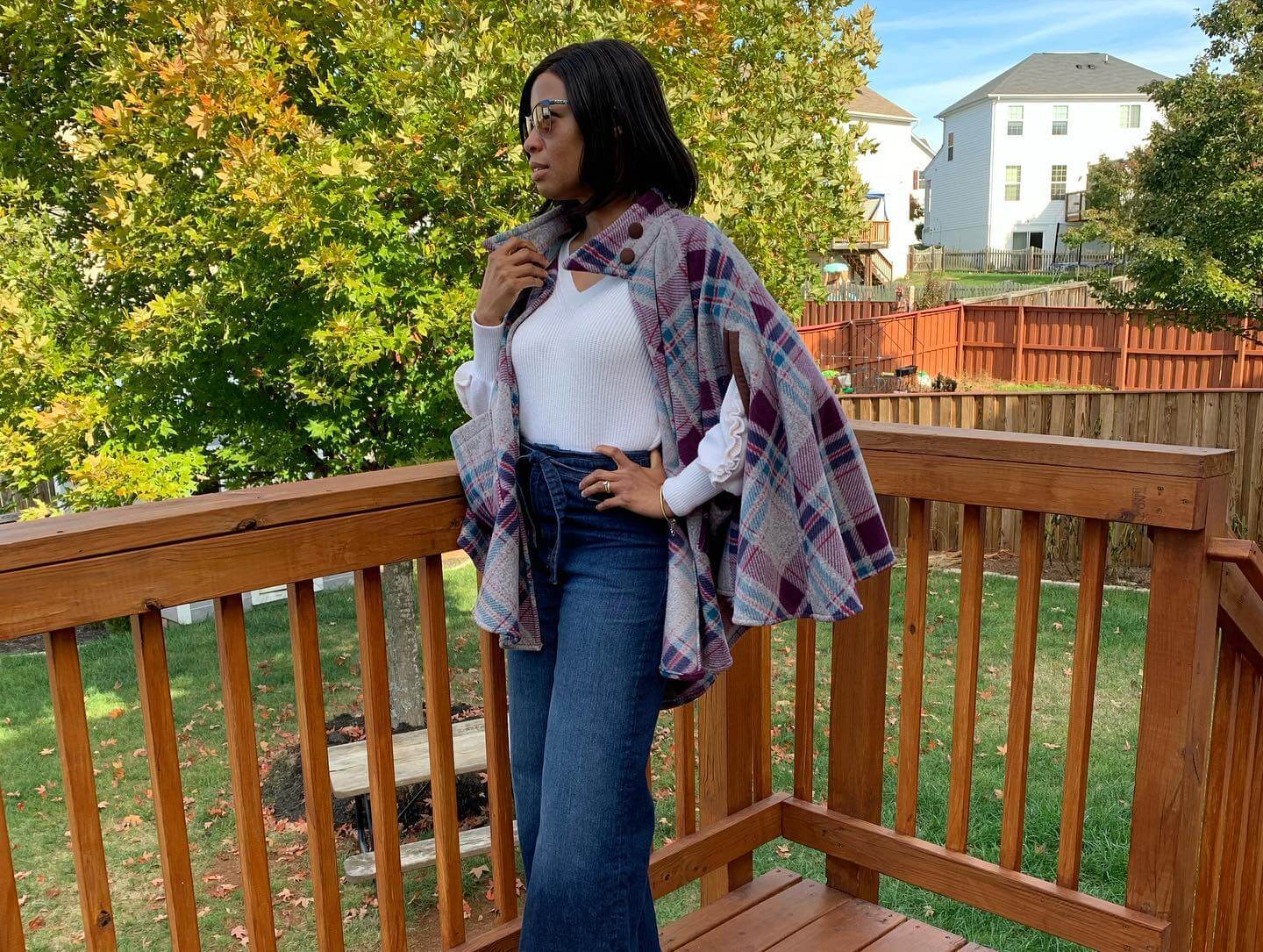 A woman stands on a wooden deck wearing a white shirt, blue jeans, and a multicolored plaid cape. She is facing sideways with one hand on her chin, looking into the distance. The background shows a wooden fence, trees with autumn foliage, and neighboring houses.
