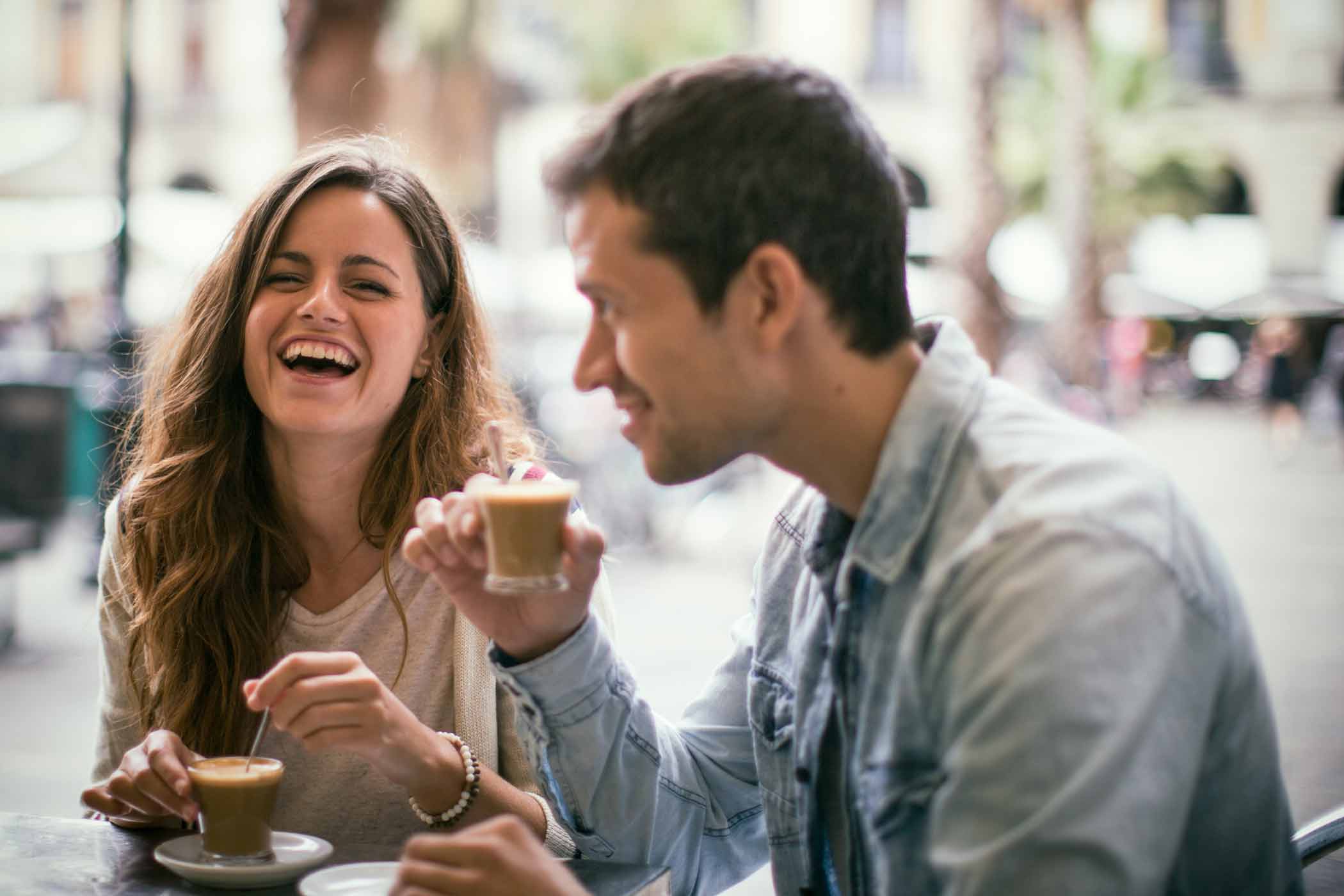 Two people are enjoying coffee at an outdoor café. The woman on the left is laughing, and the man on the right is holding a small cup of coffee and smiling. The background shows blurred outdoor scenery with trees and tables.