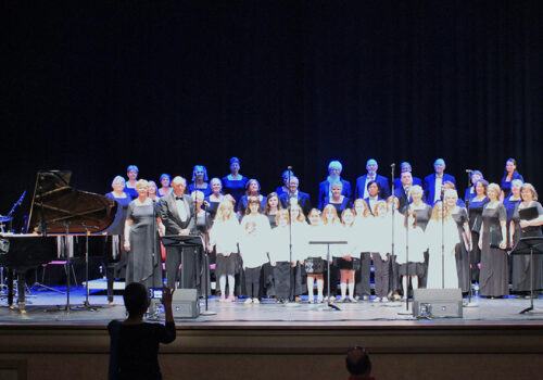 A mixed-age choir stands on stage during a performance. The front row features children in white shirts, while adults in black attire stand behind them. A pianist sits at a grand piano to the left, and a photographer captures the scene in the foreground. Image