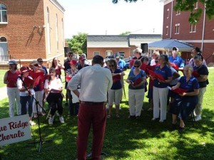 A conductor leads a choir of men and women standing on a grassy area near a brick building. Some are holding red folders. A stand with a microphone and a sign, partially readable as