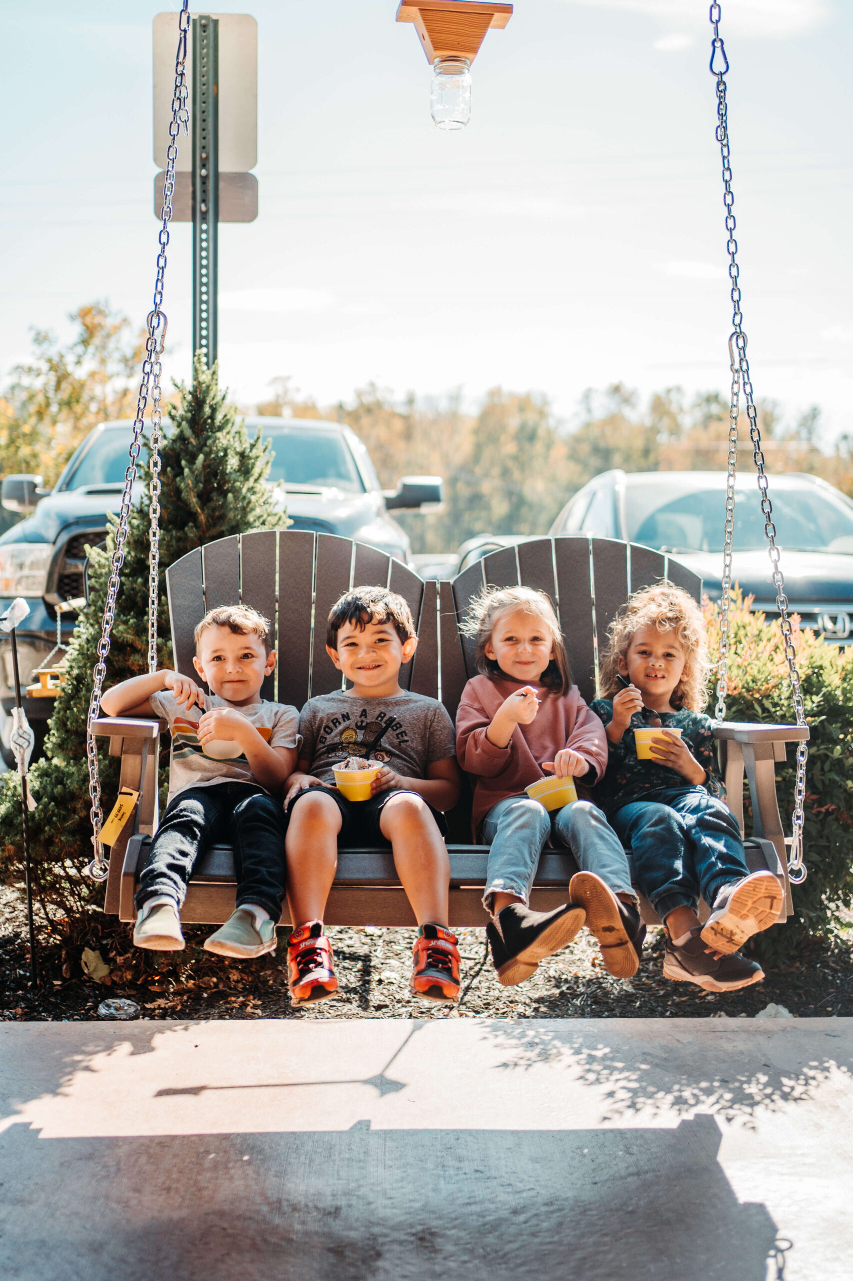 Four children sit on a wooden swing bench outdoors, smiling and eating snacks. The background includes parked cars and greenery, suggesting a relaxed, sunny day. The children look happy and engaged, enjoying their time together.
