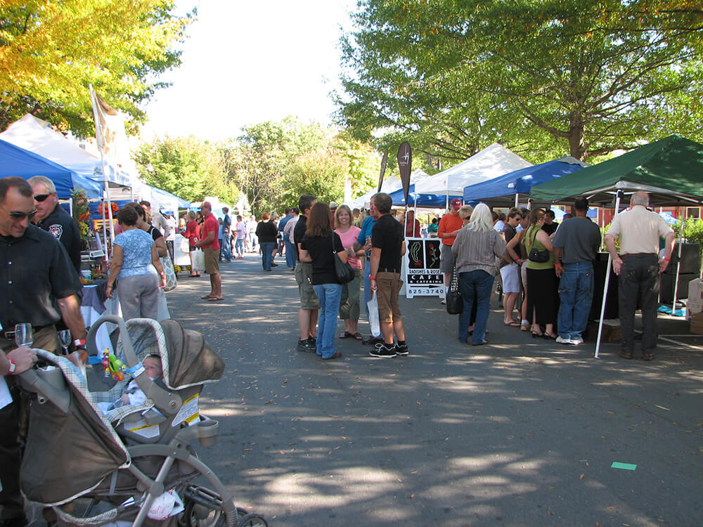 A bustling outdoor market with multiple stalls, canopies, and colorful trees. People are browsing, chatting, and mingling under the bright sun. A man pushes a stroller with a baby, and groups of visitors enjoy the lively atmosphere.