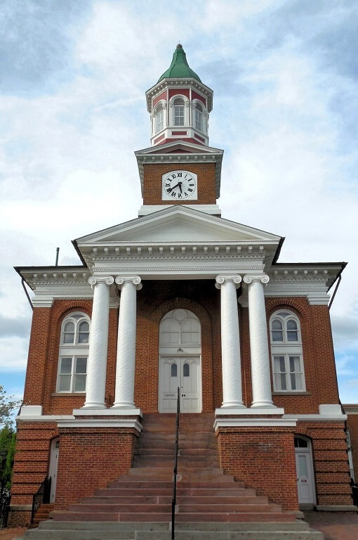 A historic brick courthouse with a clock tower and green dome. The building features four white columns at the entrance and an ornate pediment. A set of steps leads up to the main door. The sky is partly cloudy.