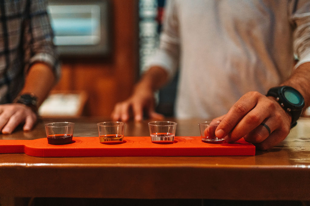 Two people are standing at a counter, each sampling a flight of drinks presented in small glasses on an orange tray. The person on the right wears a watch and a ring, with their hand resting on the tray handle.