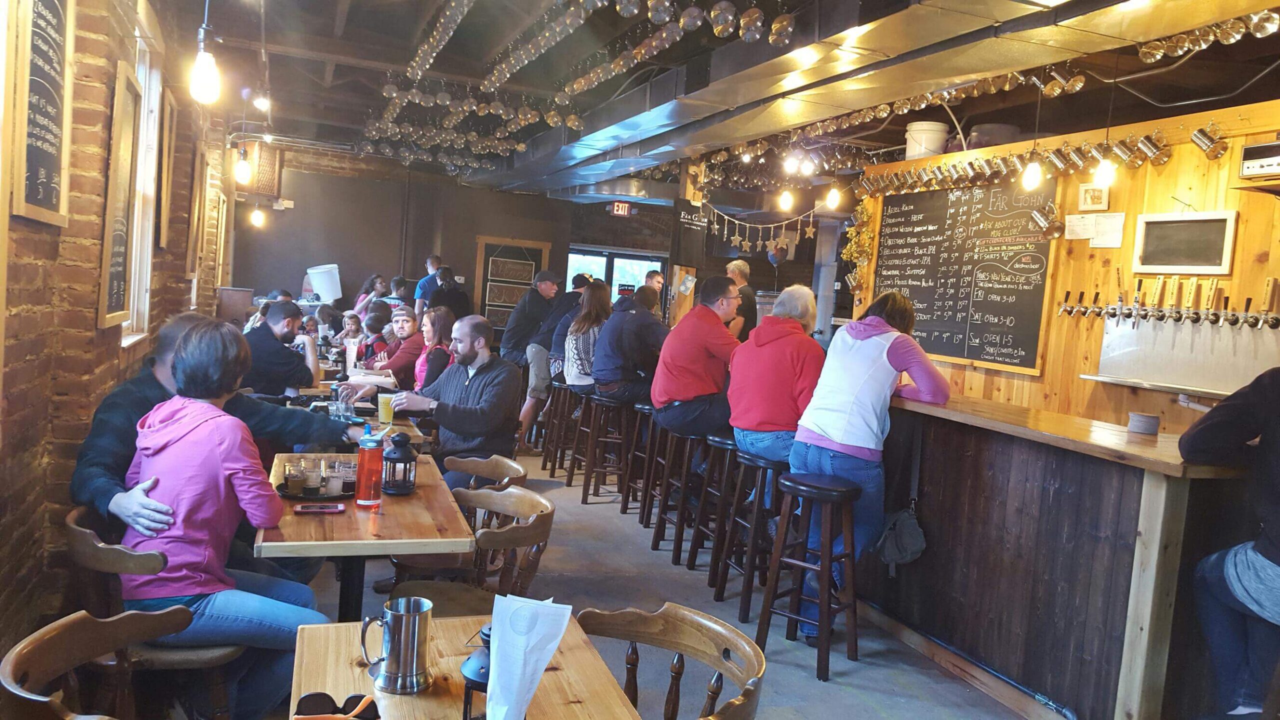 A cozy pub with patrons sitting at wooden tables and the bar. String lights hang from the ceiling, creating a warm ambiance. A chalkboard menu displays various drink options behind the bar, where several people are seated and engaged in conversation.