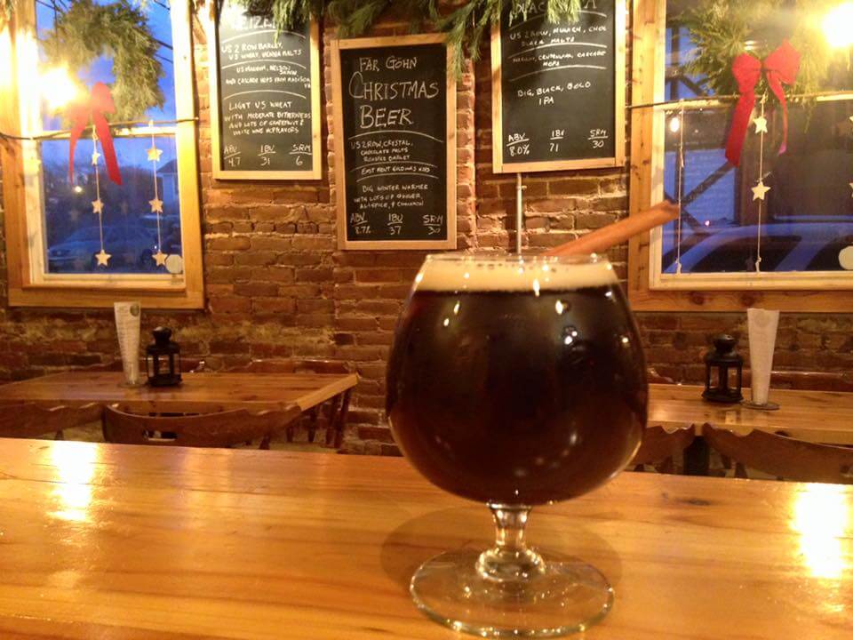 A glass of dark Christmas beer garnished with a cinnamon stick sits on a wooden bar in a cozy, warmly lit pub with brick walls. Chalkboard menus and holiday decorations can be seen in the background.