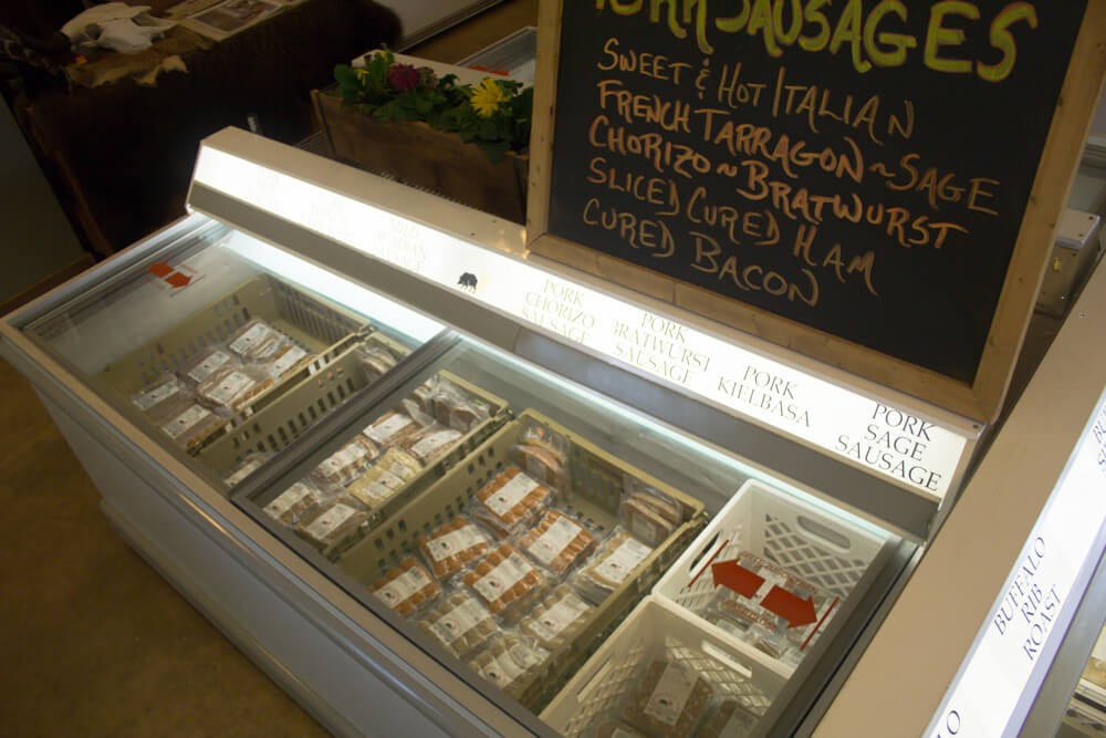 A refrigerated display case filled with various packaged meats at a market. Above the case is a chalkboard sign listing products: 