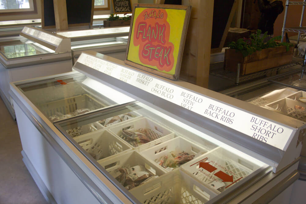 Glass-top freezers display various packages of buffalo meat, including flank steak, short ribs, and back ribs, all labeled on the freezer lids. A sign reading 