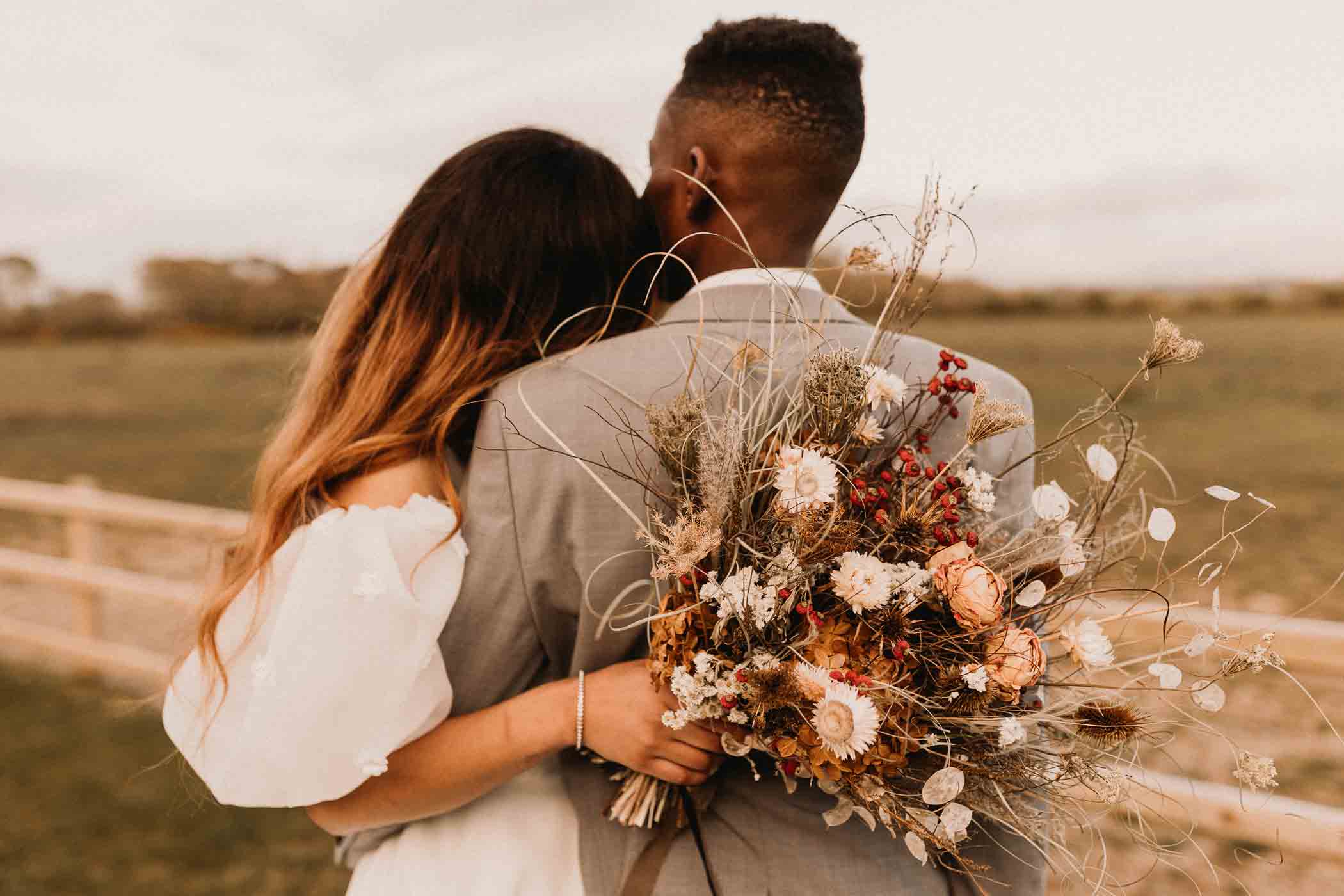 A couple stands together outdoors, facing away from the camera. The woman, in a white dress, embraces the man, who wears a gray suit. She holds a bouquet of dried and fresh flowers, including white daisies and red berries, beside a wooden fence with greenery in the background.