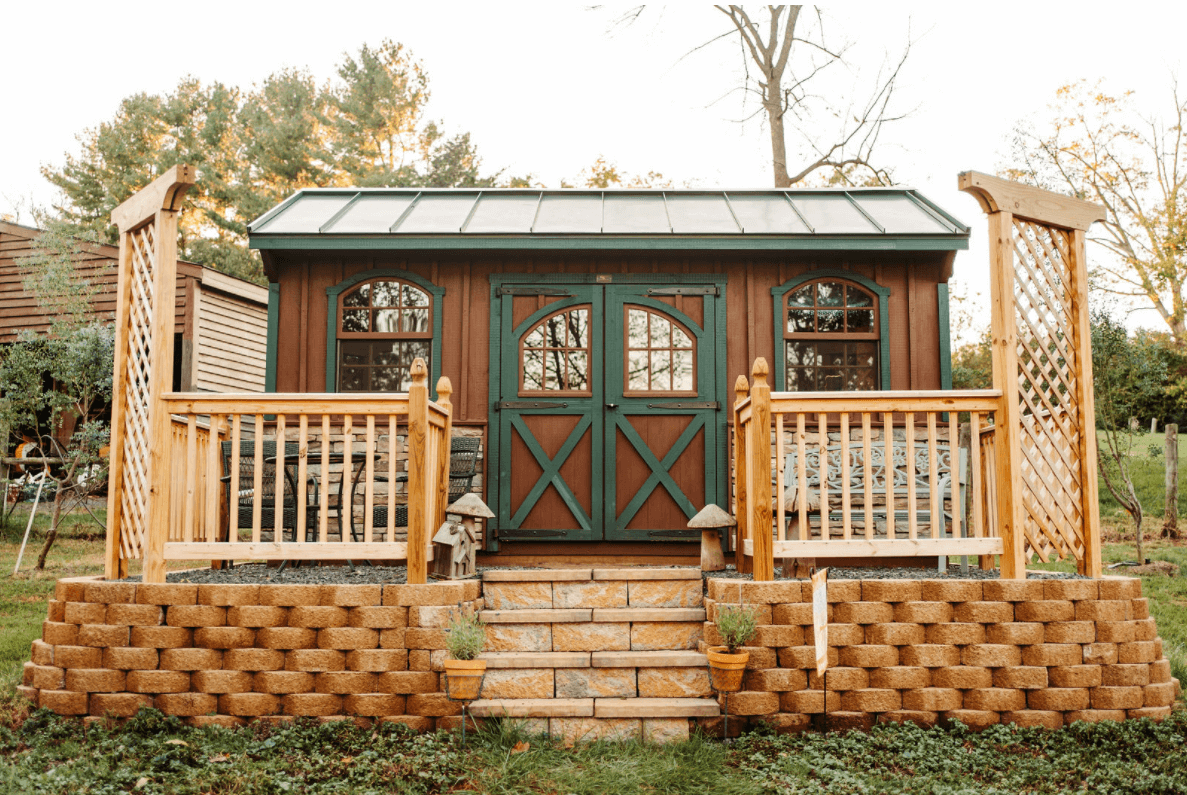 A small, charming shed with wooden double doors and arched windows, flanked by rustic railings on a raised stone platform. The platform is lined with potted plants and supported by a brick retaining wall. Trees and greenery surround the shed, creating a cozy outdoor space.