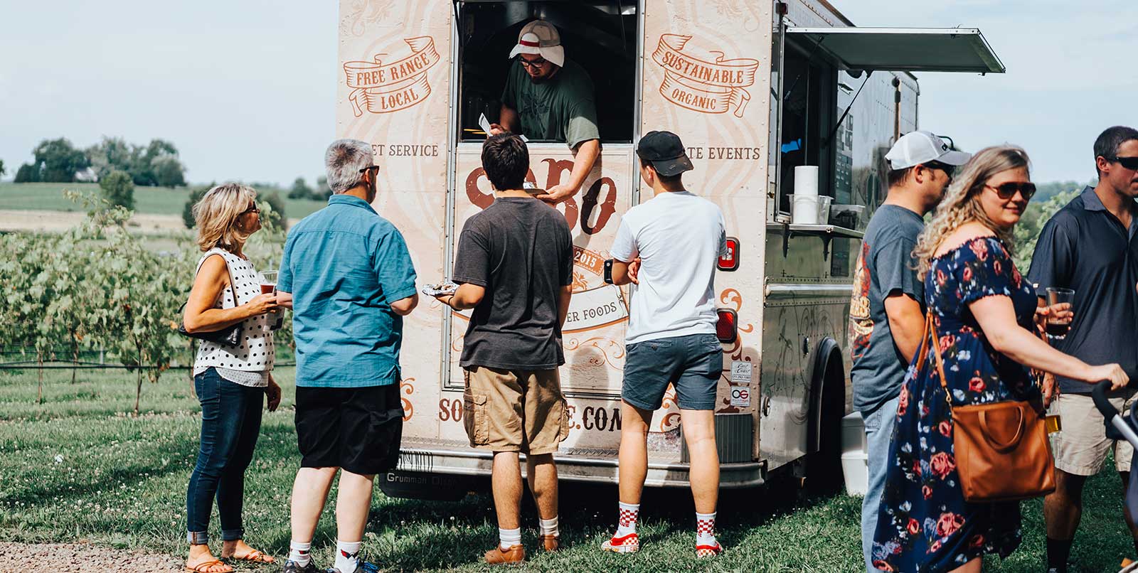 A group of people stands in line outside a food truck on a sunny day. The truck has a large sign on the back with the word 