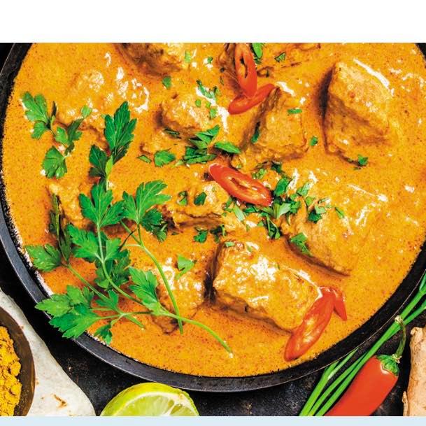 A close-up of a pan filled with vibrant orange curry featuring tender chunks of meat garnished with fresh parsley and sliced red chili peppers. Surrounding the pan are fresh herbs, spices, and a lime wedge, enhancing the dish's appealing presentation.